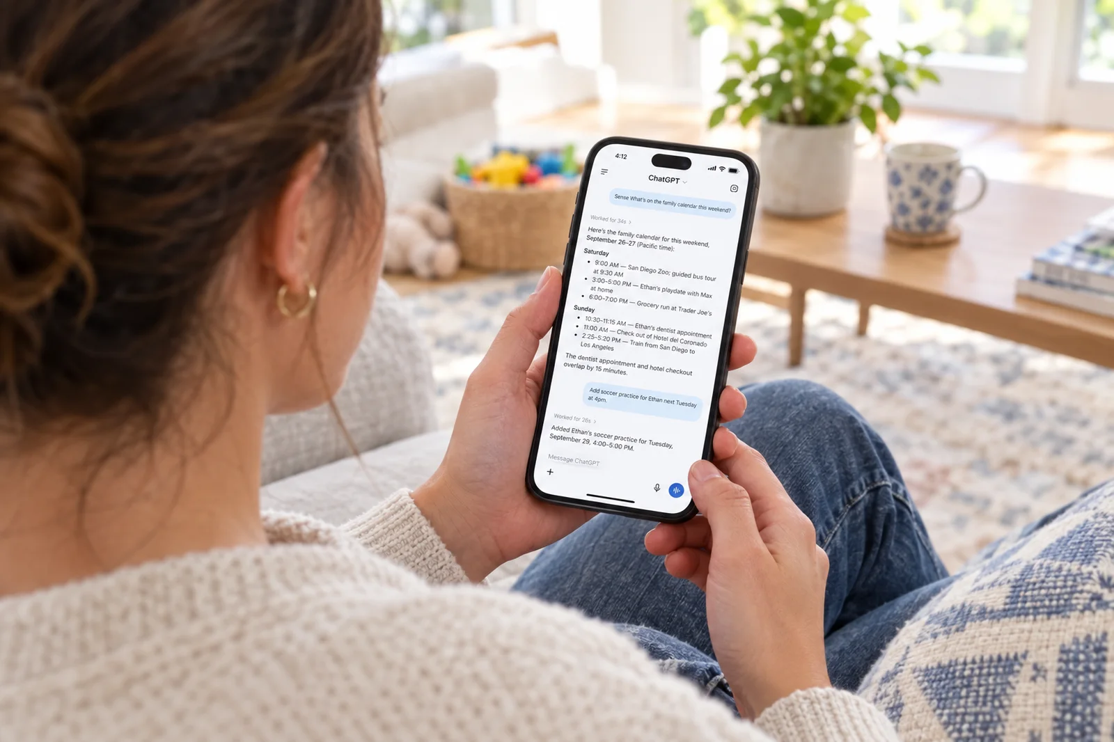 A mom on a couch reading a ChatGPT conversation on her phone in which Sense lists the family's weekend and adds soccer practice for Ethan, a toy basket and school papers nearby