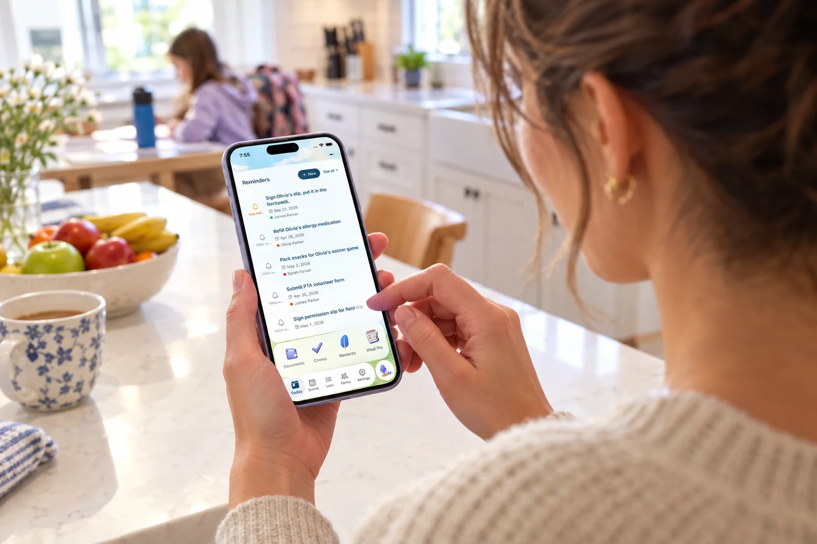 Over-the-shoulder view of a mom at a white kitchen counter reading the Sense Reminders screen on her phone, with Sign Olivia's slip, put it in the backpack at the top, while her daughter does homework at the table behind her