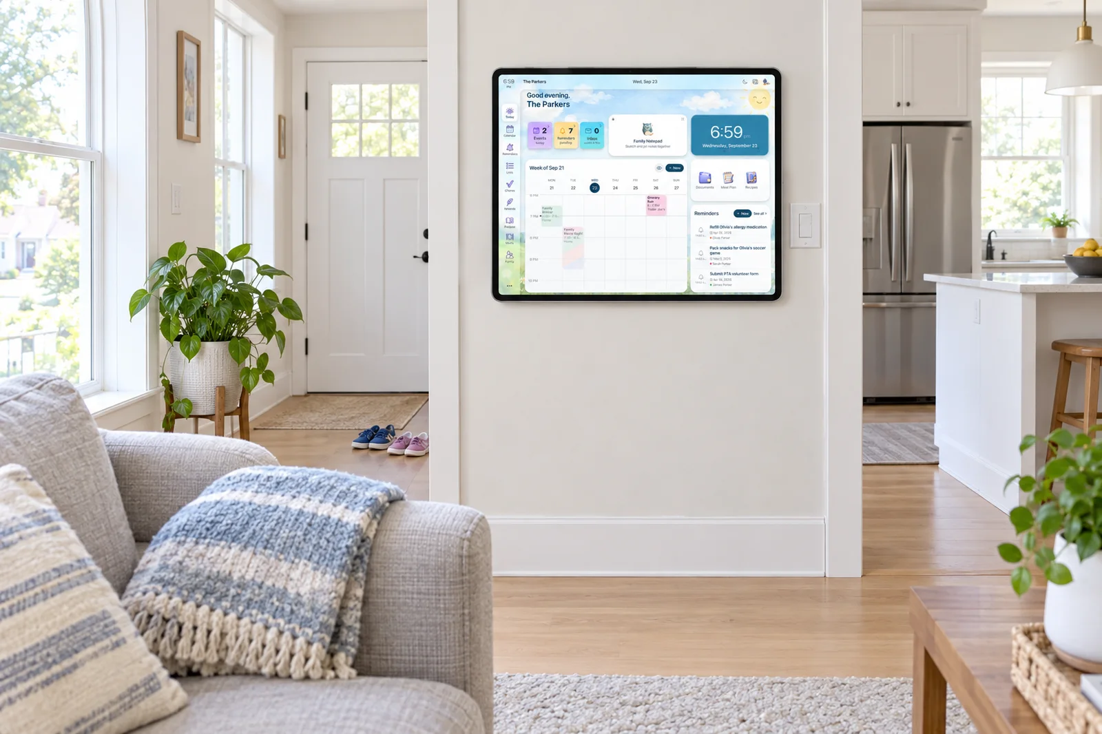 A bright living room with a tablet in a slim wall mount beside the kitchen doorway running the Sense Hub display, kids' shoes by the door
