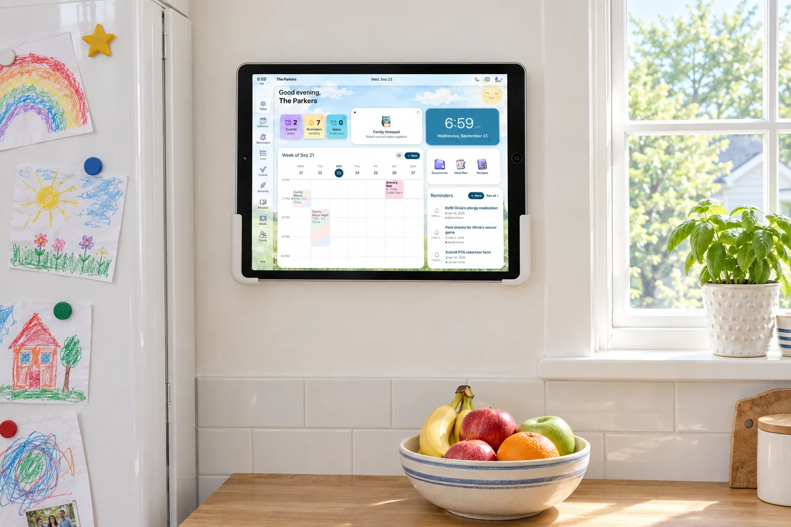 An old iPad in a plain white wall mount on a kitchen wall, running the Sense Hub family display next to a fridge covered in kids' drawings