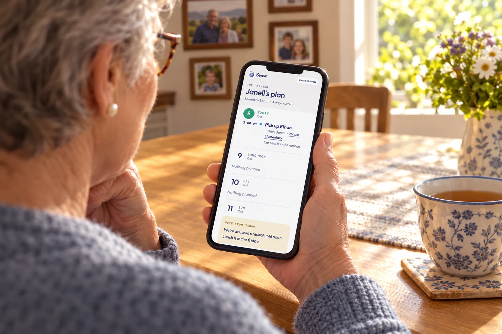A grandmother at her sunlit kitchen table with a cup of tea, reading Janell's plan on her phone: pick up Ethan at 3 and a note from Sarah about the weekend